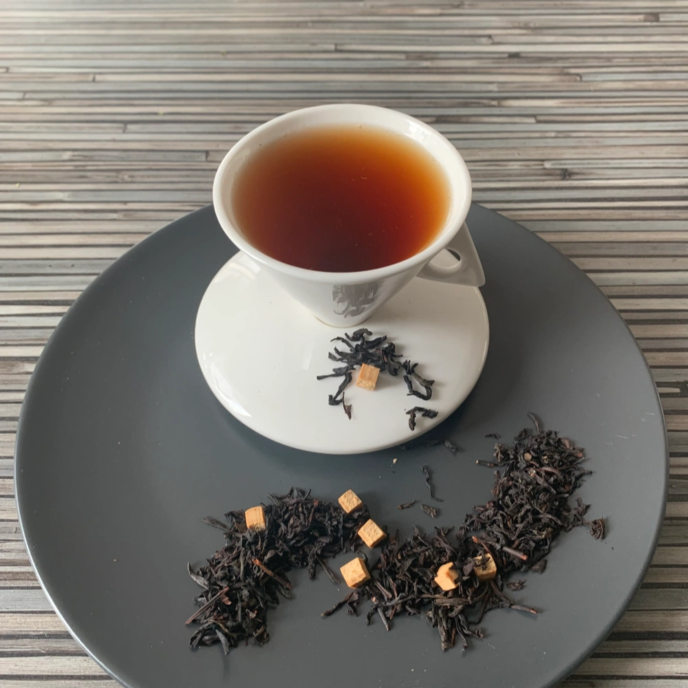 Schwarztee Karamell mit Karamellstücken in weißer Tasse auf grauem Teller vor gestreiftem Holzhintergrund