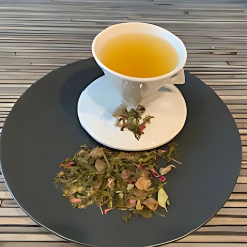 Grüner Tee Sweet Date in weißer Tasse aufgegossen, lose grüne Teeblätter mit Früchten und Blüten auf grauem Teller vor gestreiftem Holzhintergrund