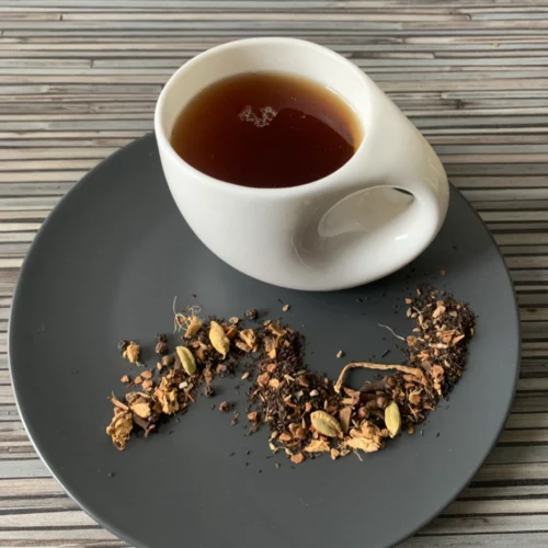 Black Chai mit Schwarztee in weißer Tasse auf dunklem Teller auf gestreifter Holzplatte, loser Tee mit Gewürzstücken daneben