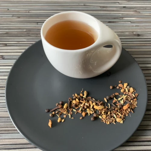 Gewürztee Herb Chai in weißer Tasse auf dunklem Teller auf gestreifter Holzplatte, loser Chai mit Gewürzen daneben