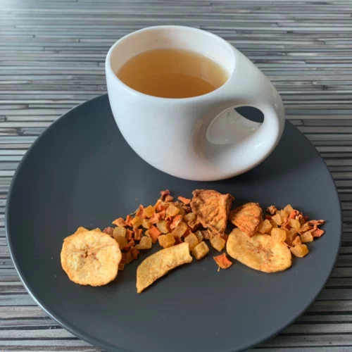 Früchtetee Anna Banana Ananas Bananen Note in weißer Tasse auf grauem Teller vor gestreiftem Holzhintergrund