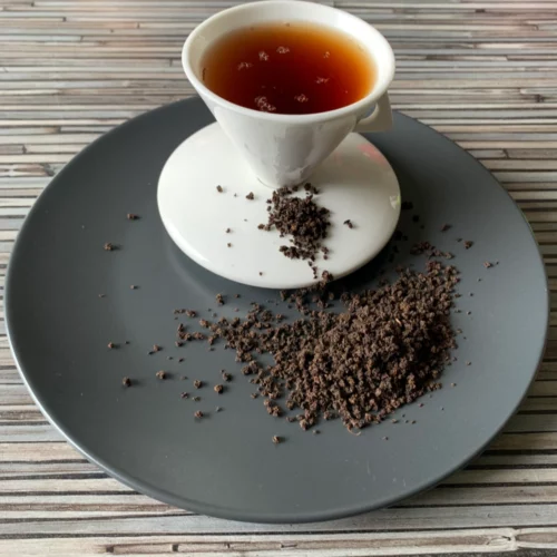 Grauer Teller mit weißer Porzellantasse dunklem Inhalt und Assam Bukhial lose dekoriert.