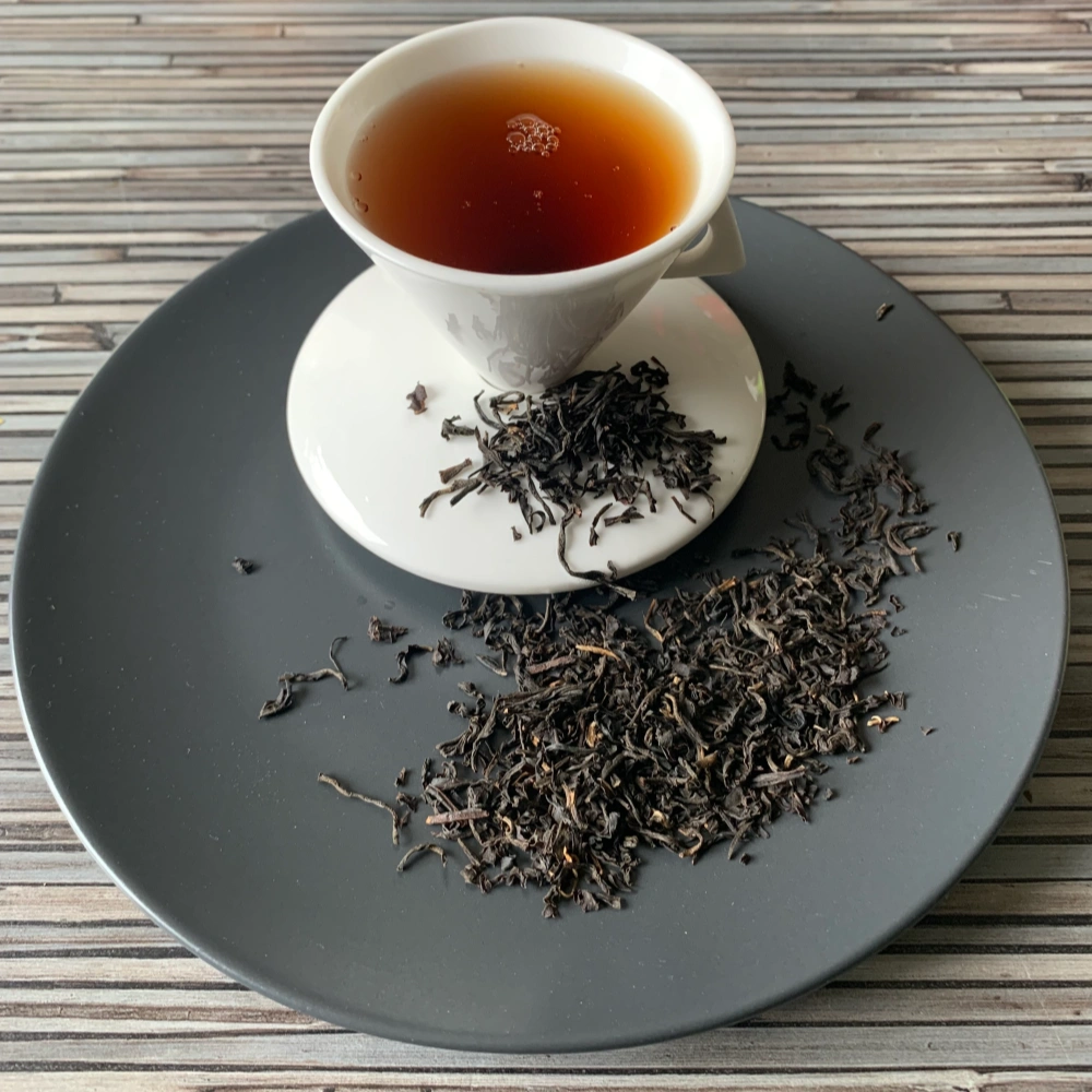 Grauer Teller mit weißer Porzellantasse dunklem Inhalt und Assam Hatidubi Smokey lose dekoriert.