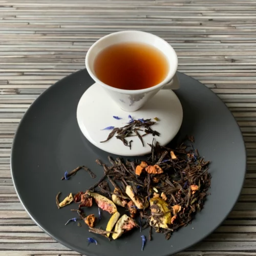 Schwarz-Grüntee Black Sencha Süße Feige in weißer Tasse auf dunklem Teller auf gestreifter Holzplatte, loser Tee mit Feigenstücken daneben