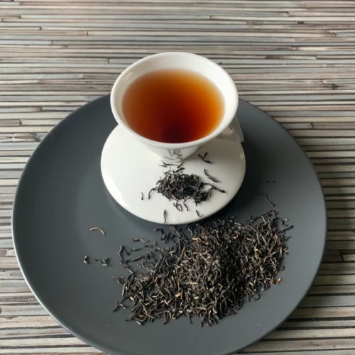 Ceylon Ratnapura FOP Special auf einem Teller auf gestreifter Holzplatte angerichtet, daneben eine weiße Tasse schwarzer Tee.