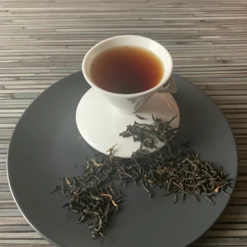 Schwarztee China Yunnan Imperial FOP auf grauem Teller mit weißer Tasse vor gestreiftem Holzhintergrund