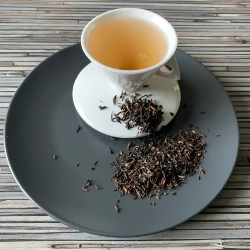 Schwarztee Darjeeling Second Flush Blattmischung auf grauem Teller mit weißer Tasse vor gestreiftem Holzhintergrund.