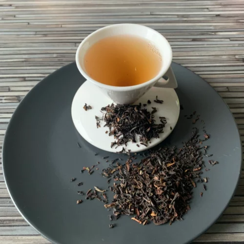 Schwarztee Darjeeling Happy Valley FTGFOP1 auf grauem Teller mit weißer Tasse vor gestreiftem Holzhintergrund