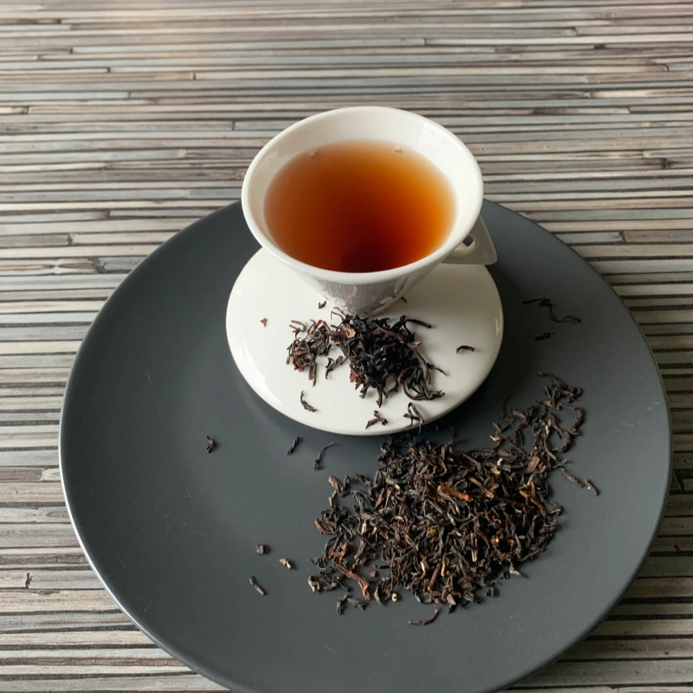 Schwarztee Darjeeling Steinthal Second Flush SFTGFOP1 auf grauem Teller mit weißer Tasse vor gestreiftem Holzhintergrund