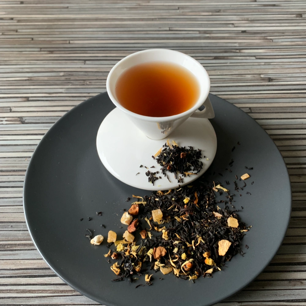 Schwarztee Ein Hauch Italien mit Blüten und Mandeln in weißer Tasse auf grauem Teller vor gestreiftem Holzhintergrund