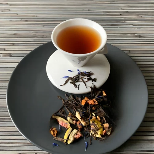 Schwarztee Feigentanz mit Feigenstücken und Kornblumenblüten in weißer Tasse auf grauem Teller vor gestreiftem Holzhintergrund