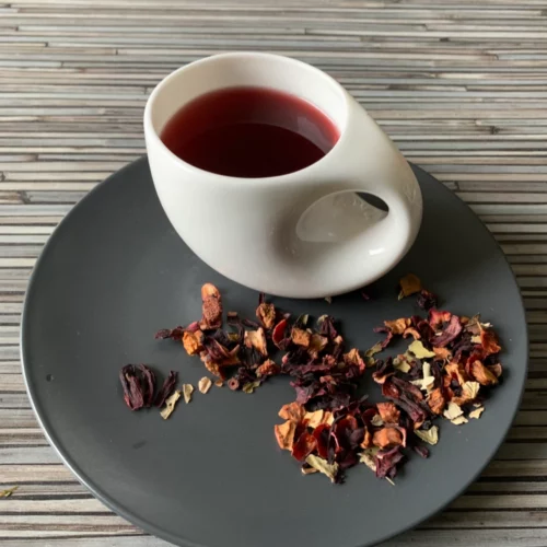 Früchtetee Alte Liebe lose auf grauem Teller, weiße Tasse mit Erdbeer‑Himbeertee auf Holzstruktur.