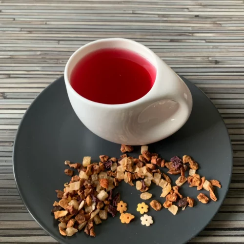 Früchtetee Mochi Flower in weißer Tasse aufgegossen und lose auf grauem Teller vor gestreiftem Holzhintergrund