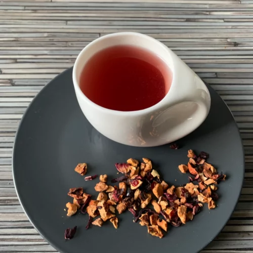 Früchtetee Sanddorn Note in weißer Tasse aufgegossen und lose auf grauem Teller vor gestreiftem Holzhintergrund