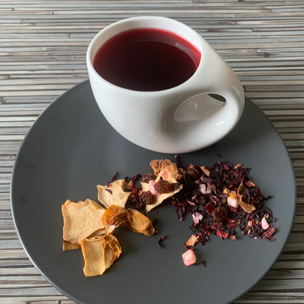 Früchtetee Sonnenfrüchte in weißer Tasse aufgegossen und lose auf grauem Teller vor gestreiftem Holzhintergrund