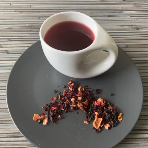 Früchtetee Wildkirsche in weißer Tasse aufgegossen und lose auf grauem Teller vor gestreiftem Holzhintergrund