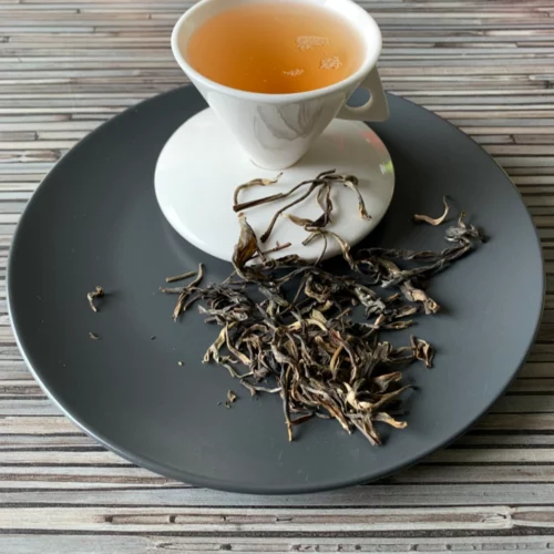 China Kekecha in weißer Tasse auf dunklem Teller auf gestreifter Holzplatte, lose gelbe Teeblätter daneben