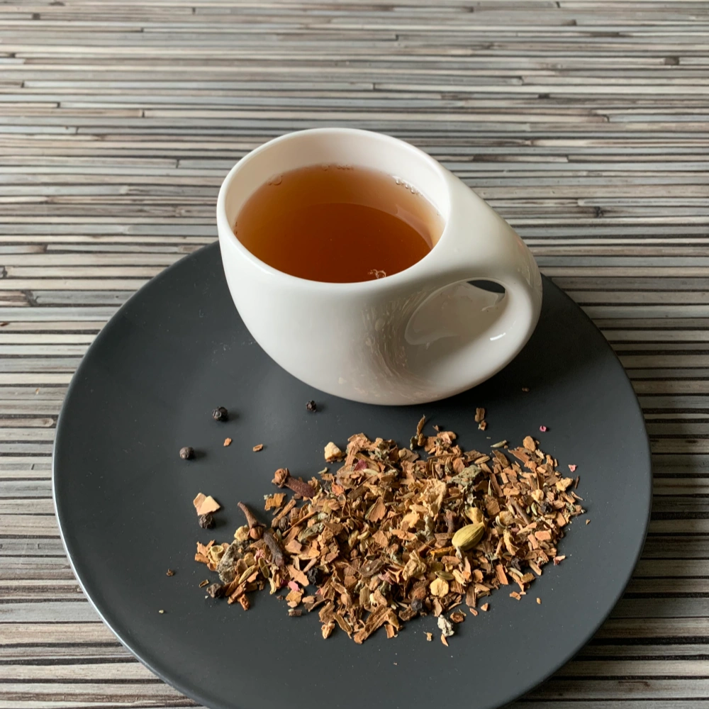 Gewürztee Chai Classic ohne Zusätze in weißer Tasse auf dunklem Teller auf gestreifter Holzplatte, loser Gewürztee mit Zimt, Kardamom und Nelken daneben