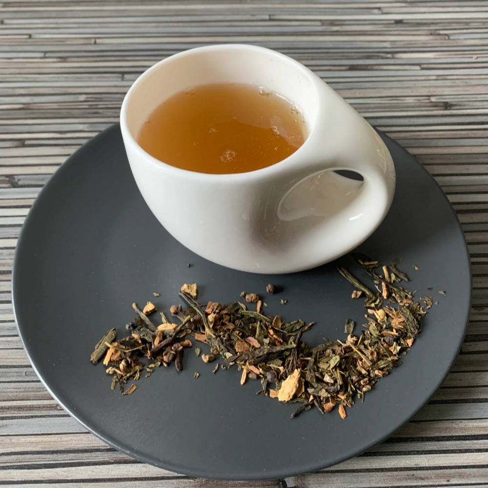 Gewürztee Green Chai mit Grüntee in weißer Tasse auf dunklem Teller auf gestreifter Holzplatte, loser Tee mit Minze, Zimt und Gewürzen daneben