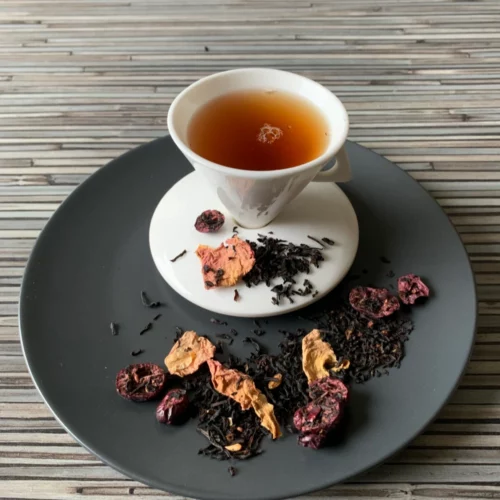 Schwarztee Granatapfel Crannyberry mit roten Fruchtstücken in weißer Tasse auf grauem Teller vor gestreiftem Holzhintergrund