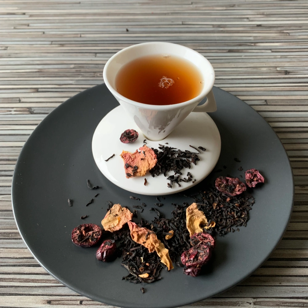 Schwarztee Granatapfel Crannyberry mit roten Fruchtstücken in weißer Tasse auf grauem Teller vor gestreiftem Holzhintergrund