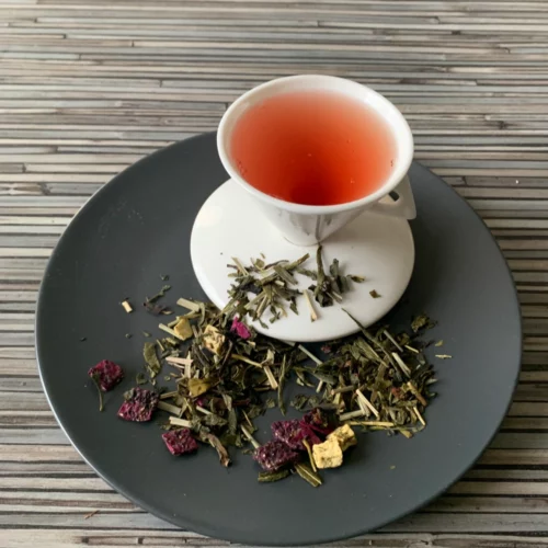 Grün-Weißer Tee Granatapfel Drachenfrucht in weißer Tasse aufgegossen, lose grüne und weiße Teeblätter mit Fruchtstücken auf grauem Teller vor gestreiftem Holzhintergrund