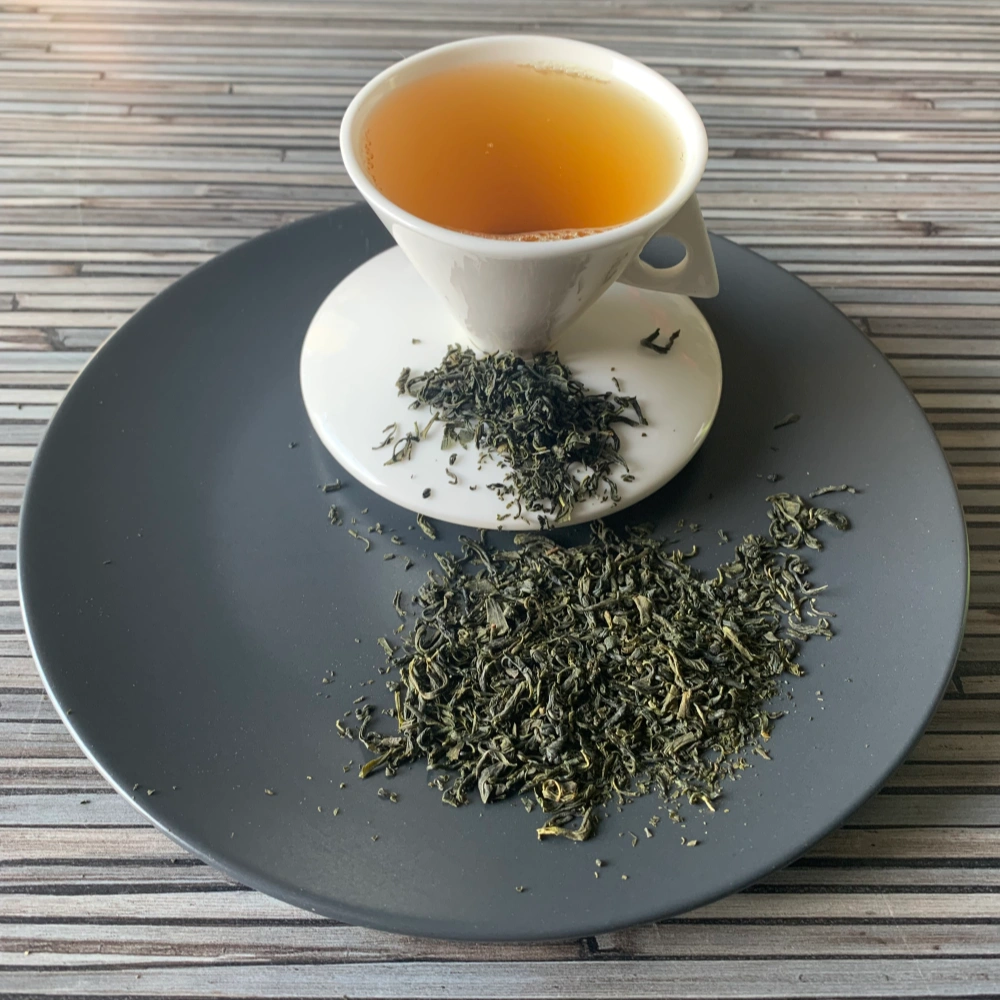 Grüner Tee Japan Tamaryokucha in weißer Tasse aufgegossen, lose Teeblätter auf grauem Teller vor gestreiftem Holzhintergrund