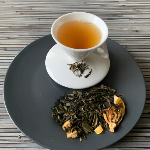 Grüner Tee Appassionato in weißer Tasse aufgegossen, lose grüne und weiße Teeblätter mit Früchten und Blüten auf grauem Teller vor gestreiftem Holzhintergrund