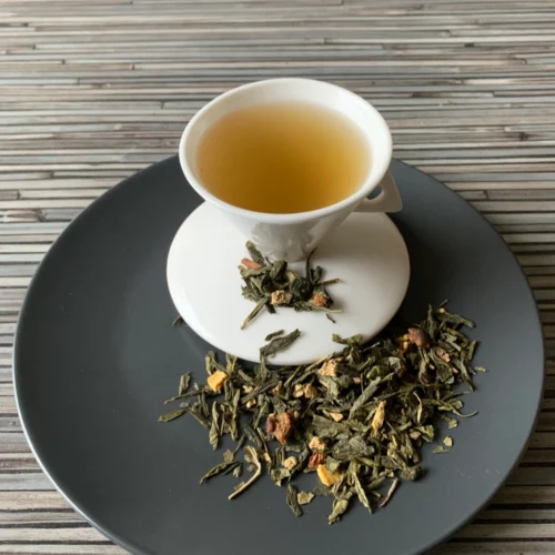 Grüner Tee Birne Ingwer Passionsfrucht in weißer Tasse aufgegossen, lose grüne Teeblätter mit Fruchtstücken auf grauem Teller vor gestreiftem Holzhintergrund