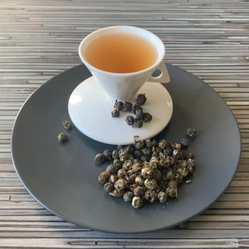 Grüner Tee China Jasmin Dragon Phoenix Pearls in weißer Tasse aufgegossen, lose Teeperlen auf grauem Teller vor gestreiftem Holzhintergrund