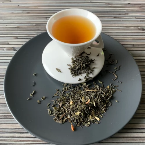 Grüner Tee China Jasmin in weißer Tasse aufgegossen, lose grüne Teeblätter mit Jasminblüten auf grauem Teller auf gestreiftem Holzhintergrund