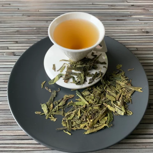 Grüner Tee China Lung Ching in weißer Tasse aufgegossen, lose Teeblätter auf grauem Teller vor gestreiftem Holzhintergrund