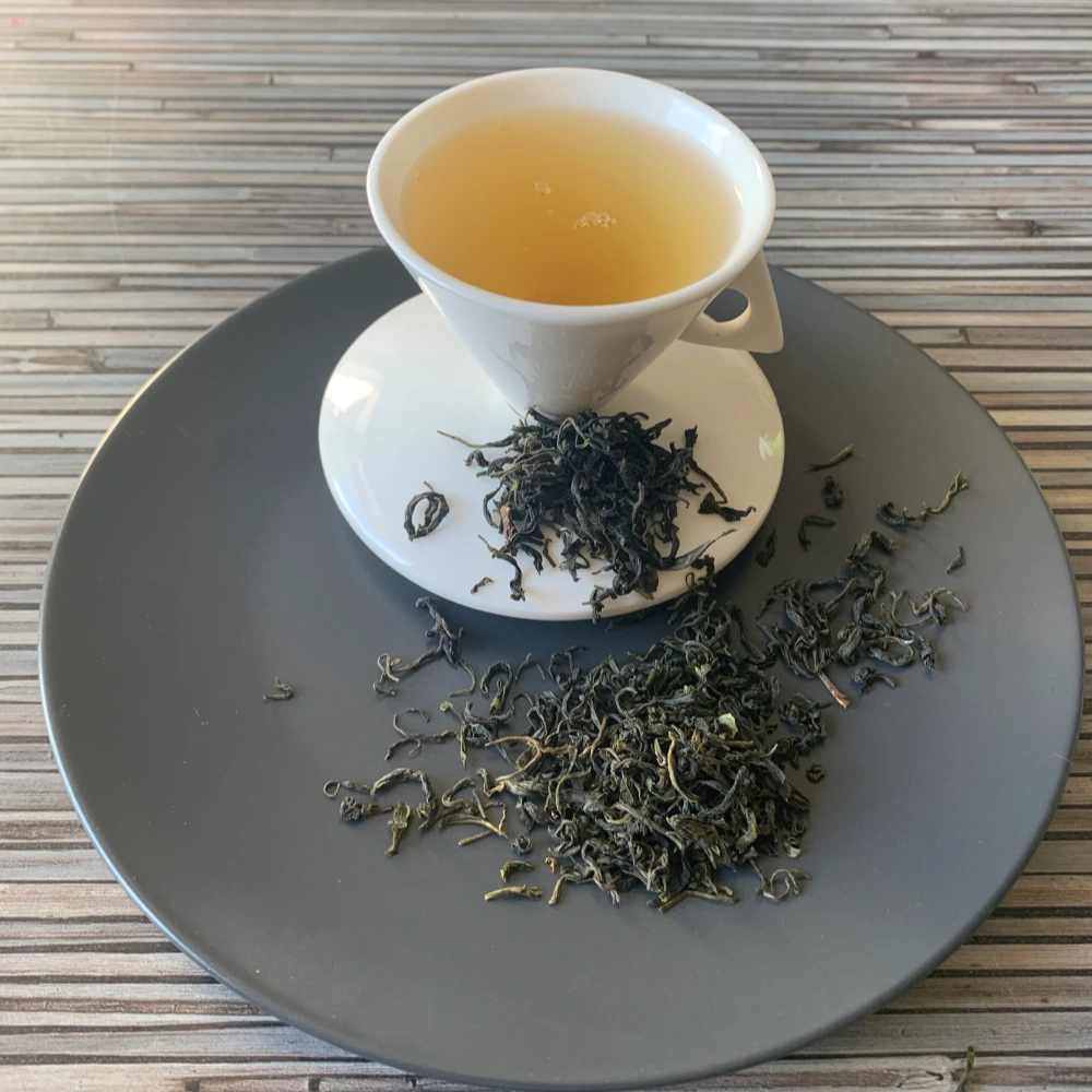 Grüner Tee China Misty Green in weißer Tasse aufgegossen, lose Teeblätter auf grauem Teller vor gestreiftem Holzhintergrund