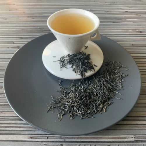 Grüner Tee China Palace Needle in weißer Tasse aufgegossen, lose Teeblätter auf grauem Teller vor gestreiftem Holzhintergrund