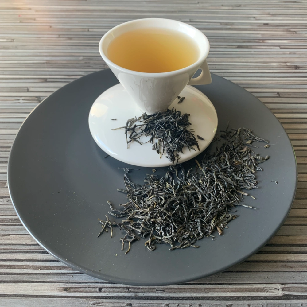 Grüner Tee China Palace Needle in weißer Tasse aufgegossen, lose Teeblätter auf grauem Teller vor gestreiftem Holzhintergrund