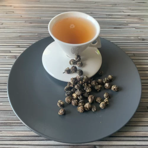 Grüner Tee China Silvery Pearl Mountain in weißer Tasse aufgegossen, lose Teeperlen auf grauem Teller vor gestreiftem Holzhintergrund