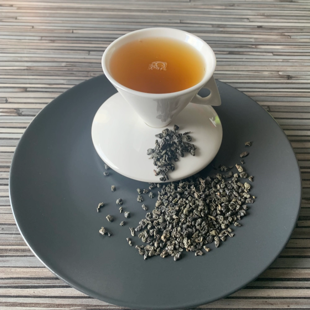 Grüner Tee China Special Gunpowder Temple of Heaven in weißer Tasse aufgegossen, lose Teeperlen auf grauem Teller vor gestreiftem Holzhintergrund