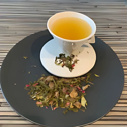 Grüner Tee Dattel Sinfonie in weißer Tasse aufgegossen, lose Grünteeblätter mit Dattelstücken, Blüten und Kräutern auf grauem Teller vor Holzhintergrund