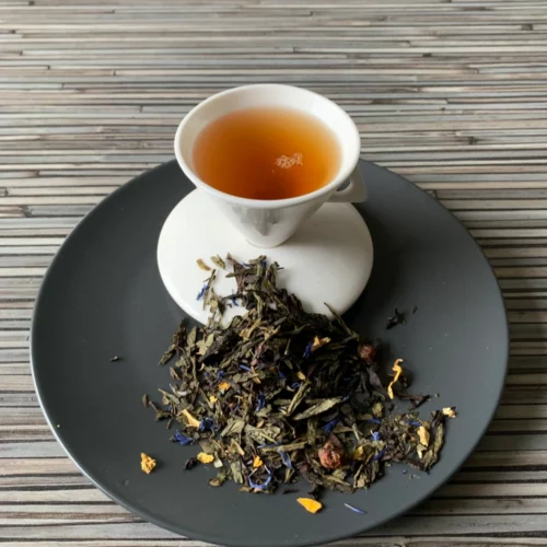 Grüner Tee Sencha Royal Star halbfermentiert in weißer Tasse aufgegossen, lose grüne Teeblätter mit Blüten auf grauem Teller vor gestreiftem Holzhintergrund