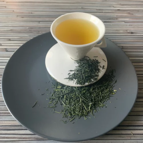 Grüner Tee Japan Gyokuro Asahi aufgegossen in heller Tasse, tiefgrüne Teeblätter auf grauem Teller vor gestreiftem Holzhintergrund