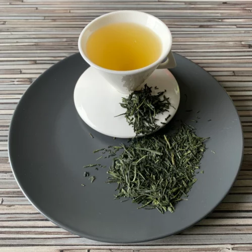 Grüner Tee Japan Sencha Fuji in weißer Tasse aufgegossen, lose Teeblätter auf grauem Teller vor gestreiftem Holzhintergrund
