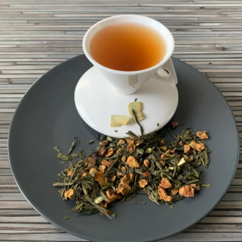 Grüner Tee Sencha Erfrischende Oase mit Matcha in weißer Tasse aufgegossen, lose grüne Teeblätter mit Kokos und Blüten auf grauem Teller vor gestreiftem Holzhintergrund