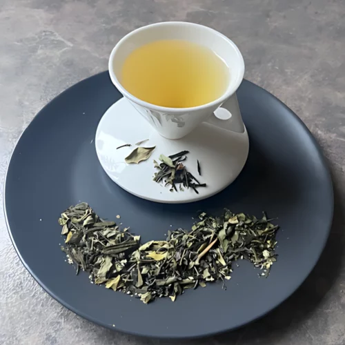 Grüntee Moringa – Leafy Green mit Moringa in weißer Tasse aufgegossen, lose grüne Teeblätter mit Fruchtstücken auf grauem Teller vor gestreiftem Holzhintergrund