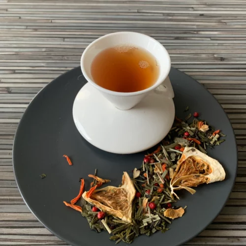 Grün-Schwarztee Kuss der Karibik in weißer Tasse aufgegossen, lose grüne und schwarze Teeblätter mit Pomelostücken, Orangenschalen und Pfeffer auf grauem Teller vor Holzhintergrund