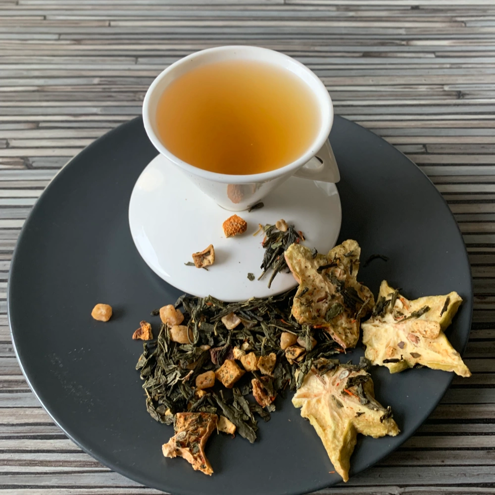 Grüner Tee Sencha Goldener Sternenstaub in weißer Tasse aufgegossen, lose grüne Teeblätter mit Fruchtstücken und Blüten auf grauem Teller vor gestreiftem Holzhintergrund