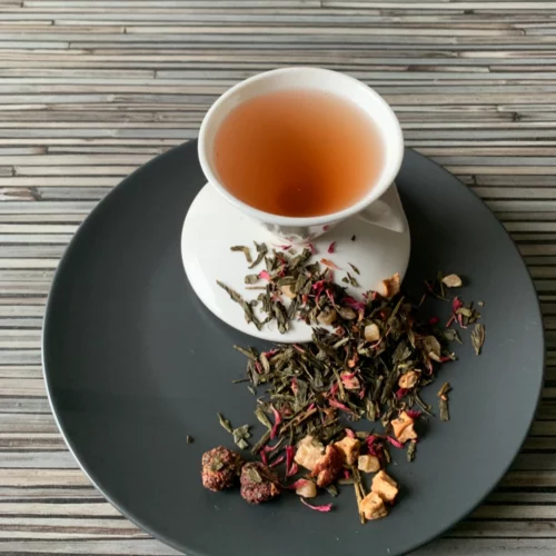 Grüner Tee Sencha Himbeertörtchen in weißer Tasse aufgegossen, lose grüne Teeblätter mit Beeren und Blüten auf grauem Teller vor gestreiftem Holzhintergrund