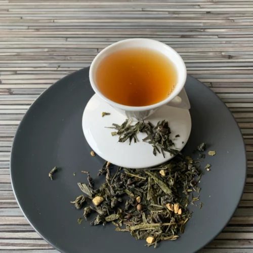 Grüner Tee Sencha Ingwer in weißer Tasse aufgegossen, lose grüne Teeblätter mit Ingwerstücken auf grauem Teller vor gestreiftem Holzhintergrund