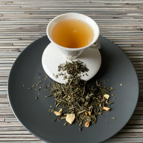 Grüner Tee Sencha Lemon in weißer Tasse aufgegossen, lose grüne Teeblätter mit Zitronenschalen auf grauem Teller vor gestreiftem Holzhintergrund