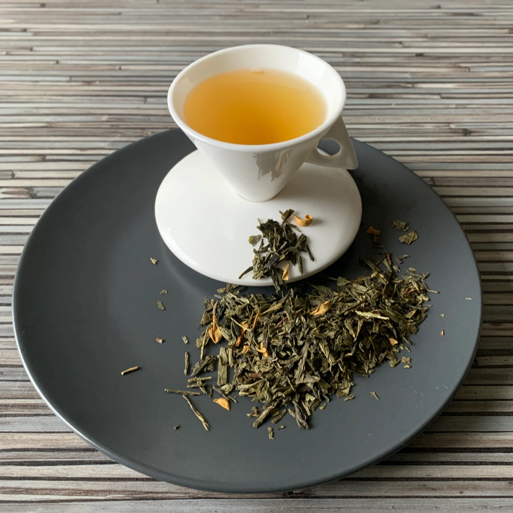Grüner Tee Sencha Orange in weißer Tasse aufgegossen, lose grüne Teeblätter mit Blüten auf grauem Teller vor gestreiftem Holzhintergrund