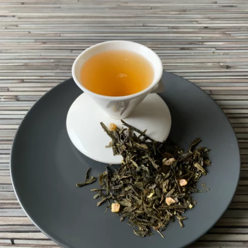 Grüner Tee Sencha Pina Colada Kokos Ananas in weißer Tasse aufgegossen, lose grüne Teeblätter mit Fruchtstücken auf grauem Teller vor gestreiftem Holzhintergrund